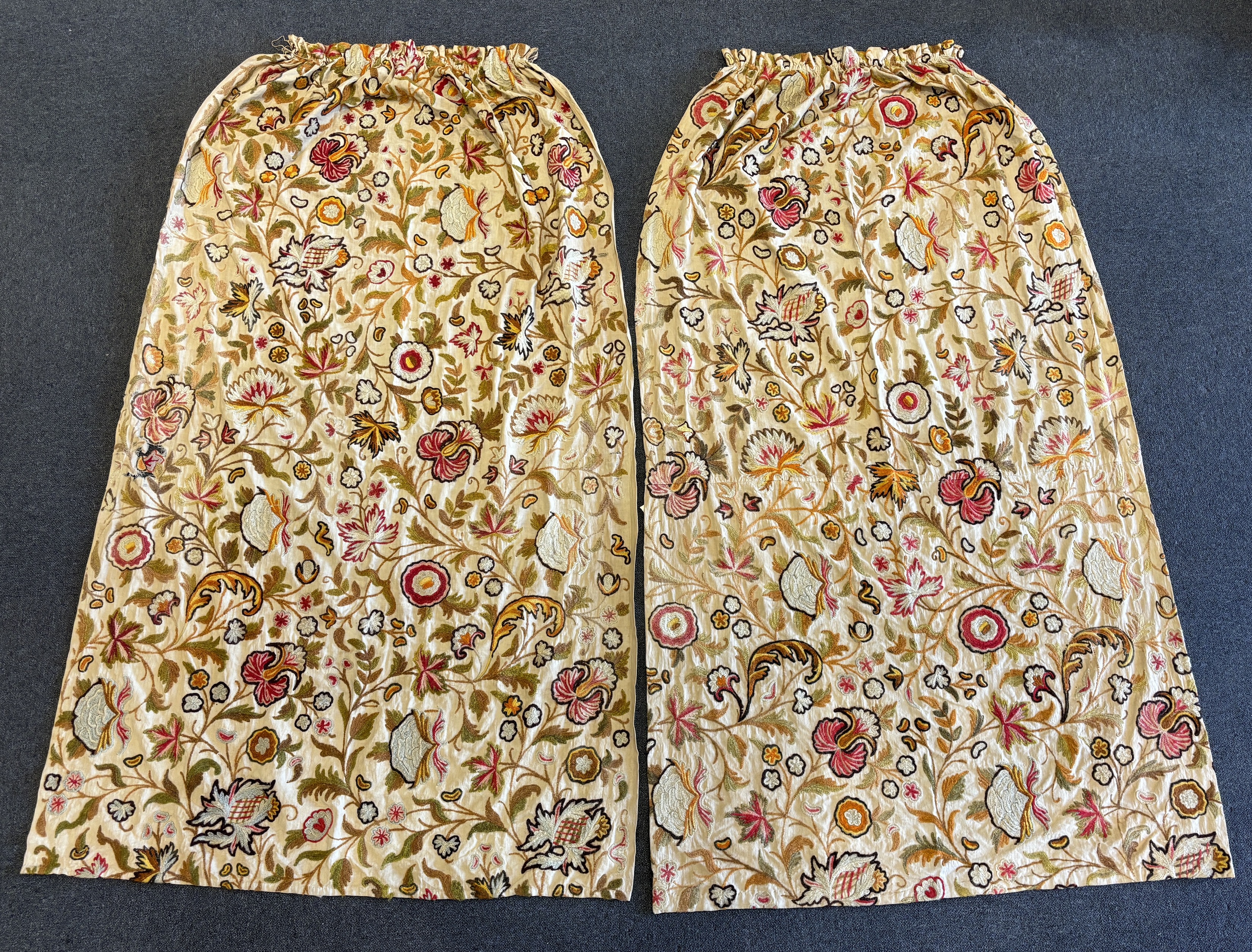 Two mid 20th century cream linen and ‘crewel work’ curtains, embroidered with chain stitch in multicoloured wools, both are unlined, one 229cm long x 116cm wide, the other 213cm long x 125cm wide